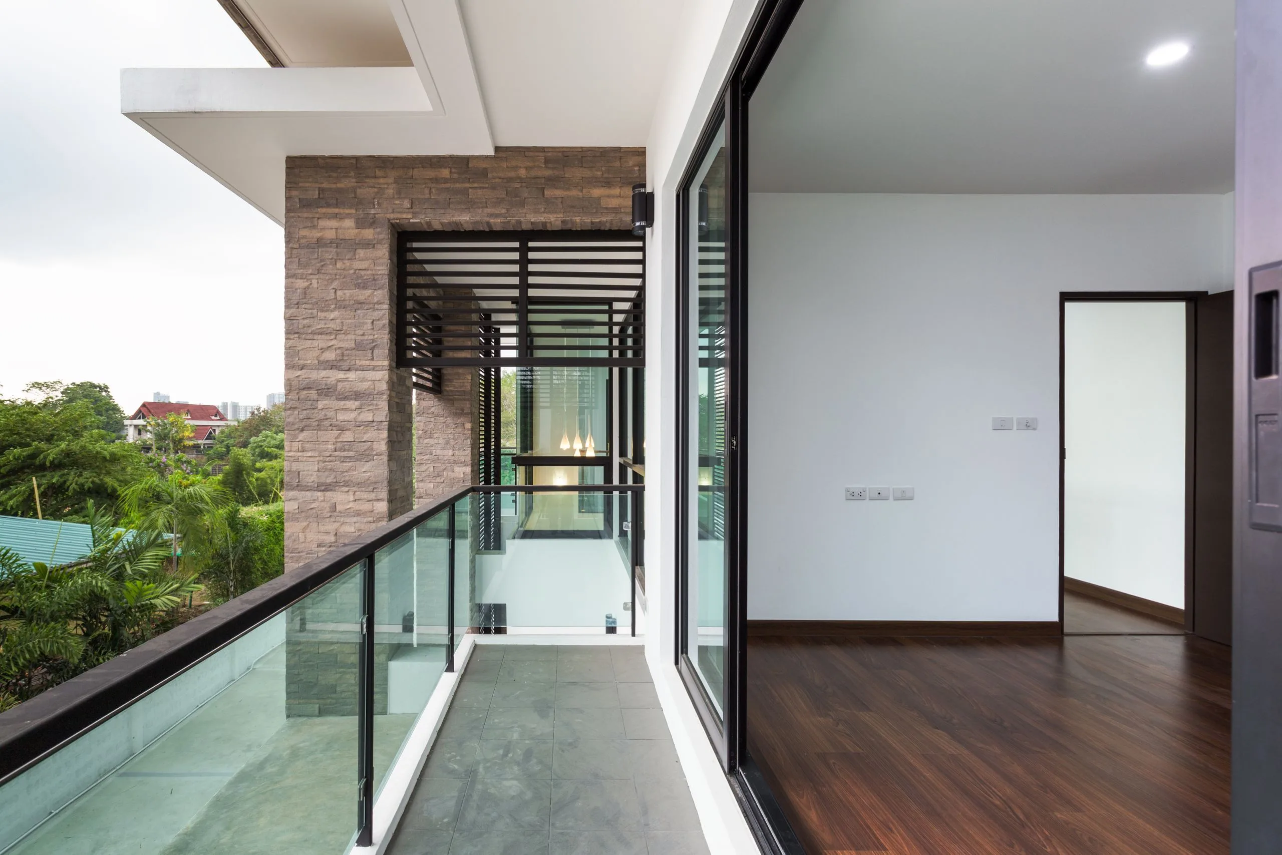 Modern apartment balcony with glass railing, adjoining room with wooden floor, and view of trees in the background.